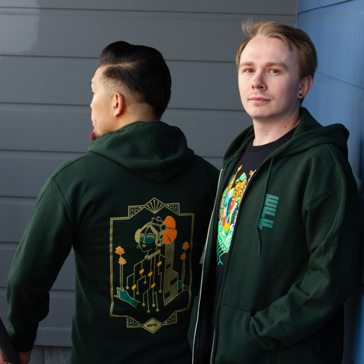 Two people wearing green hooded EFF sweatshirts with a gold "Motherboard" design on the back. Two people wearing green hooded EFF sweatshirts with a gold "Motherboard" design on the back.