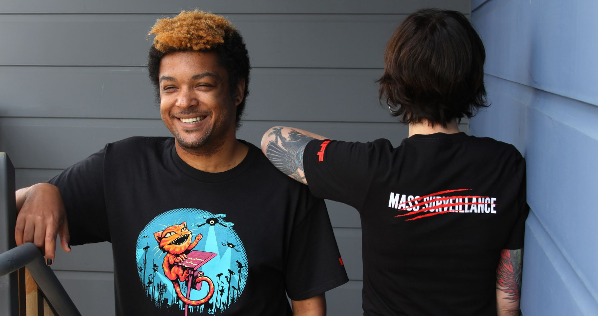 Two people wear EFF Claw Back member t-shirts. The front shows a cat swatting at spy cameras and the back says “Mass Surveillance” with red claw marks through it Two people wear EFF Claw Back member t-shirts. The front shows a cat swatting at spy cameras and the back says “Mass Surveillance” with red claw marks through it
