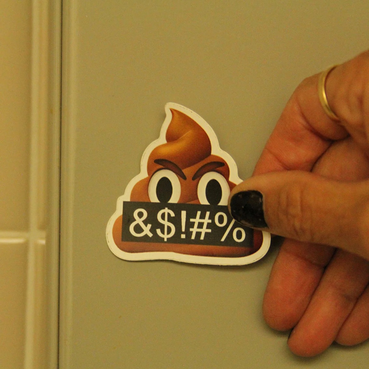 A hand with black nail polish and a gold ring holds an angry poop emoji sticker against a white door. The sticker shows a scowling poop emoji holding a black sign reading "&$!#%". A hand with black nail polish and a gold ring holds an angry poop emoji sticker against a white door. The sticker shows a scowling poop emoji holding a black sign reading "&$!#%".